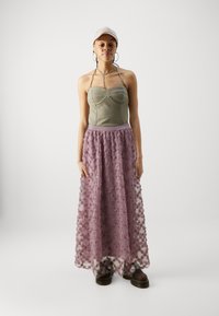 Licht olijfgroene strapless top met een getailleerde lijfje, gecombineerd met een lange, doorschijnende mauve rok met bloemenborduursel. Bruine platform schoenen.