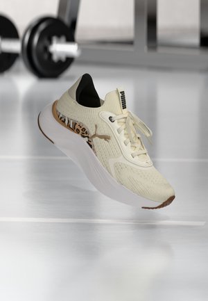Chaussure de course beige Puma avec semelle blanche et détail imprimé léopard, flottant au-dessus d’un sol de salle de sport avec des poids flous en arrière-plan.
