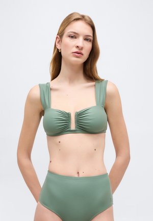 Mladá žena s dlouhými blond vlasy nosící zelené bikiny s vysokým pasem, širokými ramínky a předním průstřihem na klíč.