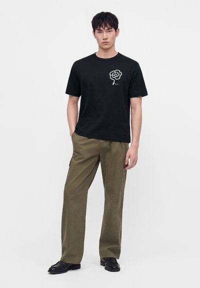 Jeune homme debout les mains dans les poches, portant un t-shirt noir à motif graphique, un pantalon cargo vert olive et des chaussures noires sur un fond clair uni.