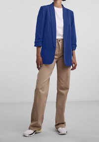 Blå blazer med uppvikta ärmar, bärs över en vit t-shirt. Kombineras med beige vida byxor och vita sneakers. Slät tygfinish.