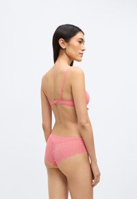 Rausvo nėrinių liemenėlė ir derančios kelnaitės; pasižymi sudėtingais geometriniais raštais, banguotais kraštais ir reguliuojamomis petnešėlėmis patogumui.