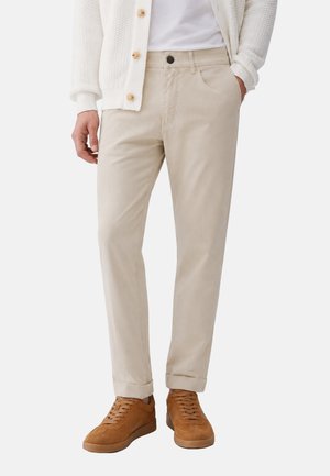 Uomo che indossa chino beige, cardigan bianco a trama, camicia bianca e sneakers in suede marrone, in piedi con una mano in tasca.