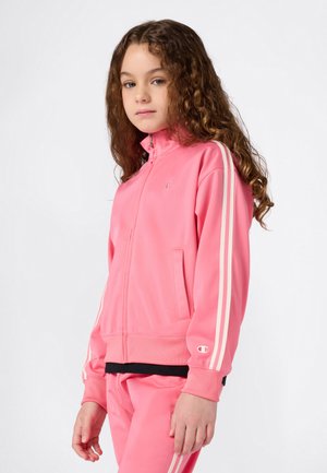 Jeune personne avec de longs cheveux bouclés portant une veste zippée rose et un pantalon assorti avec des rayures blanches sur les manches et les jambes.