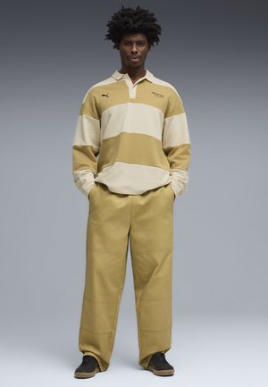 Man staat met handen in zijn zakken, draagt een beige en crème gestreept poloshirt met lange mouwen en bijpassende beige brede broekspijpen met zwarte schoenen.