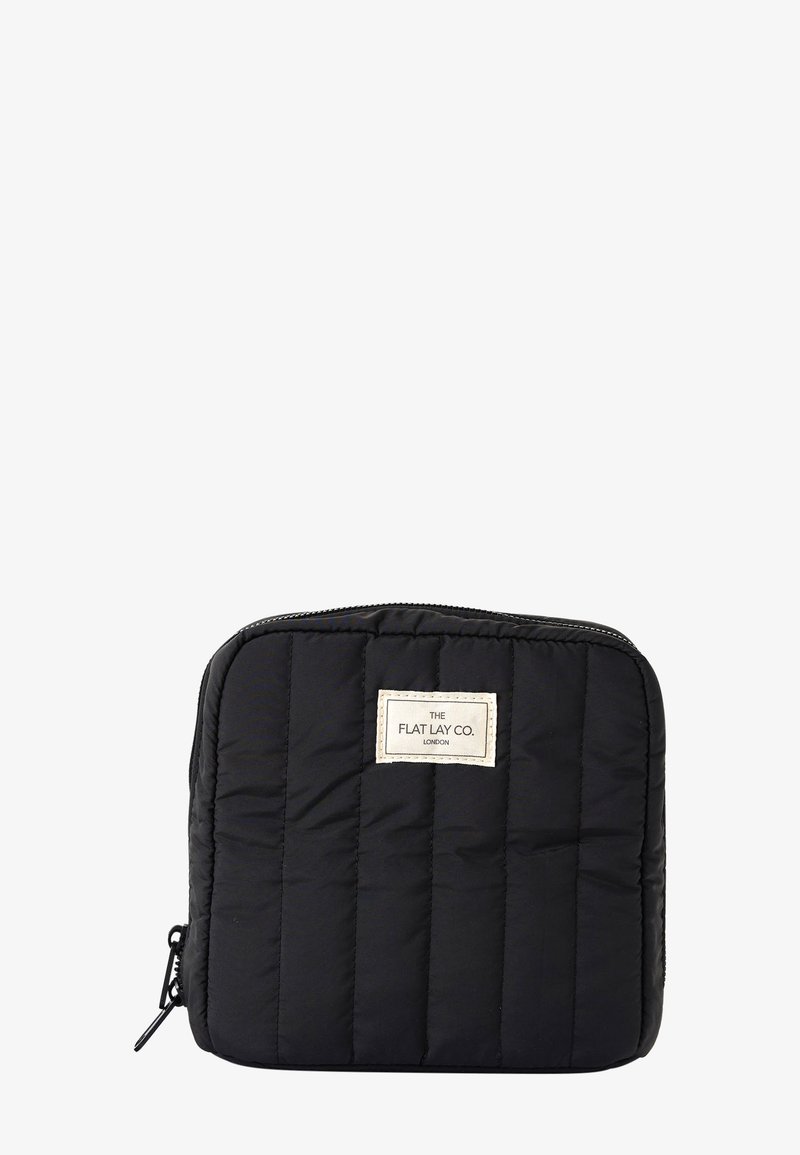 Czarna, pikowana, prostokątna saszetka z zamkiem błyskawicznym i białą prostokątną etykietą z napisem „THE FLAT LAY CO. LONDON” umieszczoną na środku z przodu.