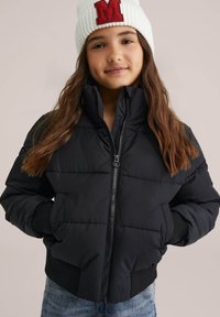 Jeune personne avec de longs cheveux portant un bonnet en tricot blanc avec un "M" rouge et une veste matelassée noire, les mains dans les poches.