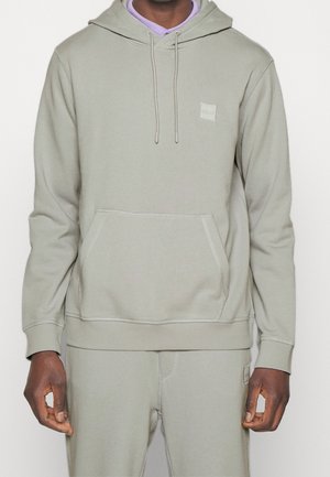 Homme portant un sweat à capuche gris clair et un pantalon de survêtement assorti avec une poche kangourou et des détails de coutures visibles.