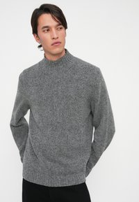 Mann trägt einen eng anliegenden grauen Strickpullover mit einem Stehkragen und langen Ärmeln und blickt seitlich vor einem weißen Hintergrund.