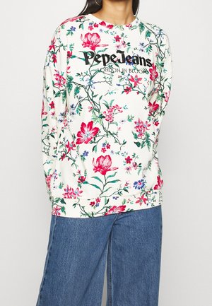Kobieta w białej, kwiatowej bluzie Pepe Jeans i niebieskich dżinsach, stojąca na jednolitym tle.