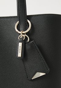 Sac fourre-tout noir texturé avec un accent en métal doré et une étiquette porte-clés gravée "GUESS". Comprend un détail triangulaire argenté.