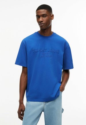 Młody mężczyzna w niebieskiej koszulce "Tommy Jeans" i jasnoniebieskich dżinsach, stojący z jedną ręką w kieszeni na jednolitym białym tle.