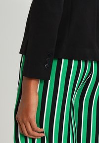 Svart blazer med en skräddarsydd design och tre blå knappar vid ärmarna, kombinerad med gröna, svarta och vita randiga byxor.