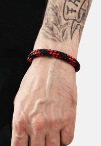 Braccialetto intrecciato nero e rosso realizzato in materiale tessuto, con una forma cilindrica che si avvolge attorno al polso. Dettagli di superficie texture visibili.