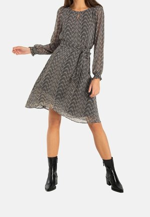 Vrouw die een lange jurk met zwarte en witte zigzagprint draagt met doorschijnende mouwen en zwarte laarzen met hak tot halverwege de kuit, poserend met gespreide benen.