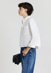 Personne portant une chemise blanche à manches longues et à boutons, un jean bleu, tenant une pochette noire en matériau souple, faisant face à gauche sur un fond neutre.