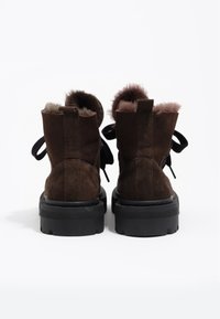 Botines de ante marrón con un suave forro de piel, suelas de goma negras y detalles de cinta de tela en la parte trasera.