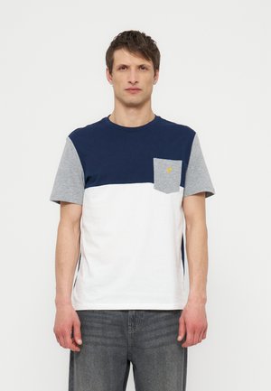 Mann står iført en fargeblokkert t-skjorte med marineblå, grå ermer, hvit nedre del og en grå brystlomme med en liten gul logo.