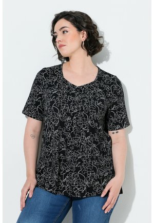 Kvinde med kort mørkt hår iført en sort top med blomsterprint og blå jeans, står op ad en lys væg og kigger mod venstre.