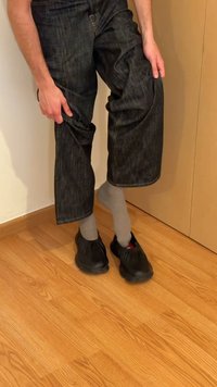 Schwarze Slipper mit elastischen Öffnungen, kombiniert mit grauen Socken. Die Person trägt dunkelblaue Jeans, die leicht hochgekrempelt sind, wodurch die Schuhe sichtbar werden.