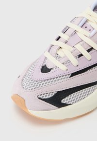 Närbild av en lila, svart och vit sneaker med krämfärgade snören och detalj i nätmaterial på en vit bakgrund.