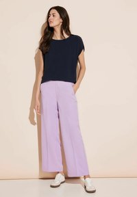 Marinblå kortärmad topp parat med vida lavendelfärgade byxor med sidofickor och vita sneakers med ljusbrun sula.