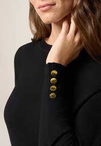 Schwarzer Pullover mit Rundhalsausschnitt, softer Textur und vier goldenen Knöpfen am linken Bündchen. Keine sichtbaren Muster oder zusätzlichen Verzierungen.
