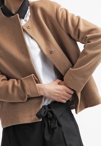 Brązowa kurtka z mieszanki wełny, z okrągłym dekoltem, zapinana na pojedynczy guzik, z długimi rękawami. Połączona z czarną spódnicą z wiązaniem w talii i białą bluzką.