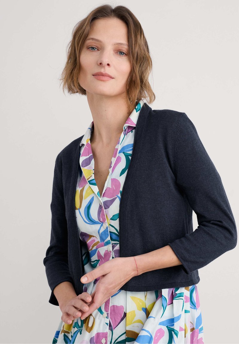 Navigično modra pletena jopica s tremi četrtinami rokavov, oblečena preko pisane cvetne obleke z gumbi na sprednji strani in ovratnikom.