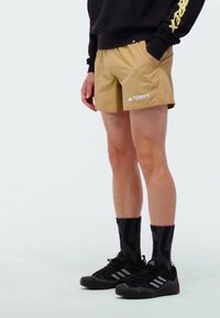 Svijetlosmeđi šortsevi s logom s lijeve strane, upareni s crnim čarapama s geometrijskim uzorkom i crnim sportskim cipelama.
