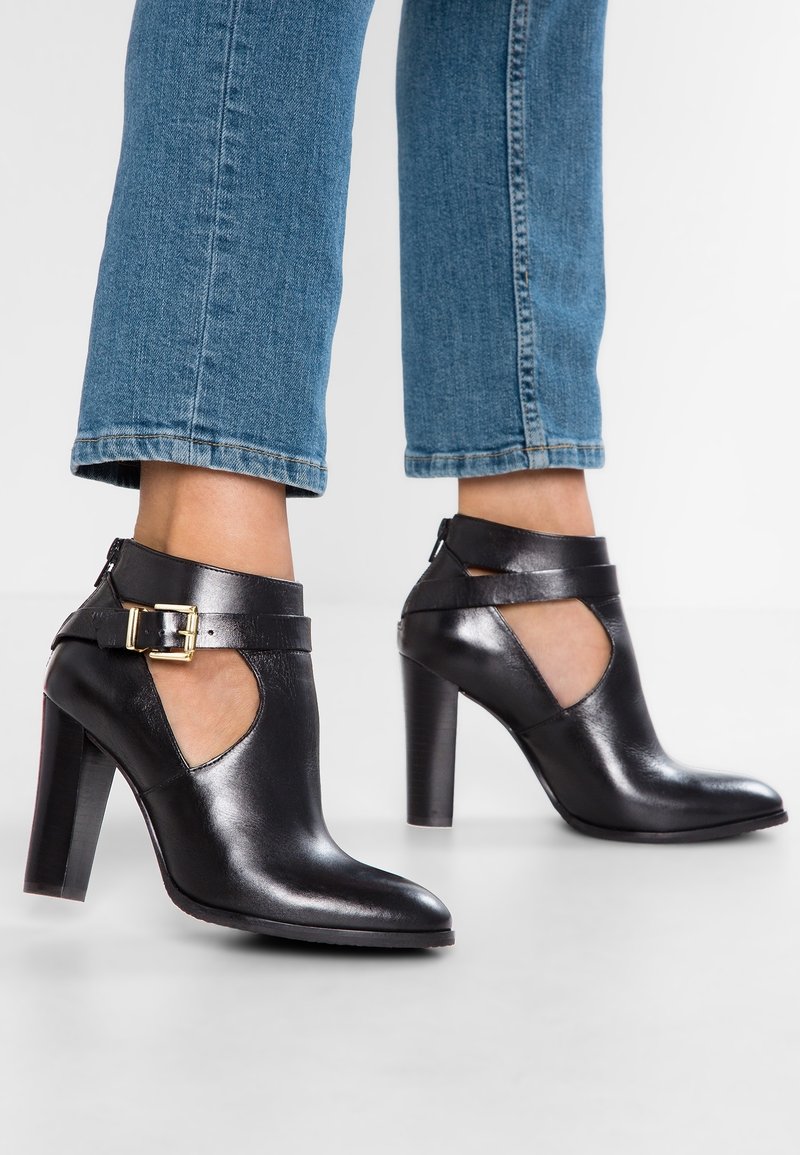 Mustat nahkaiset nilkkurit korolla, joissa on leikattu muotoilu, säädettävä solkihihna ja sileä pinta, yhdistettyinä lyhennettyihin sinisiin farkkuihin.