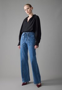 Svart langermet bluse kombinert med høy midje blå denim bukser med snøring og vide ben, supplert med burgunder hæler.