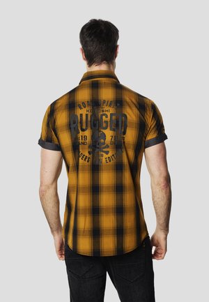 Lühikeste varrukatega ruuduline särk kollases ja mustas, millel on seljal graafiline trükk "RUGGED" ja kolju. Pööratud varrukaotsad ja lõdvestunud istuvus.