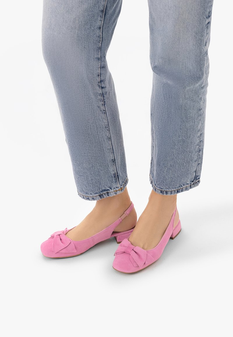 Pieds portant un jean bleu clair et des chaussures slingback roses à talon bas avec un détail nœud, debout sur un fond blanc.