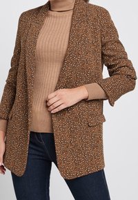 Bruine geblokte blazer met een gestructureerde afwerking, met lichte en donkere vlekken, en een klassiek notch lapel. Draagt over een geribbelde beige top.