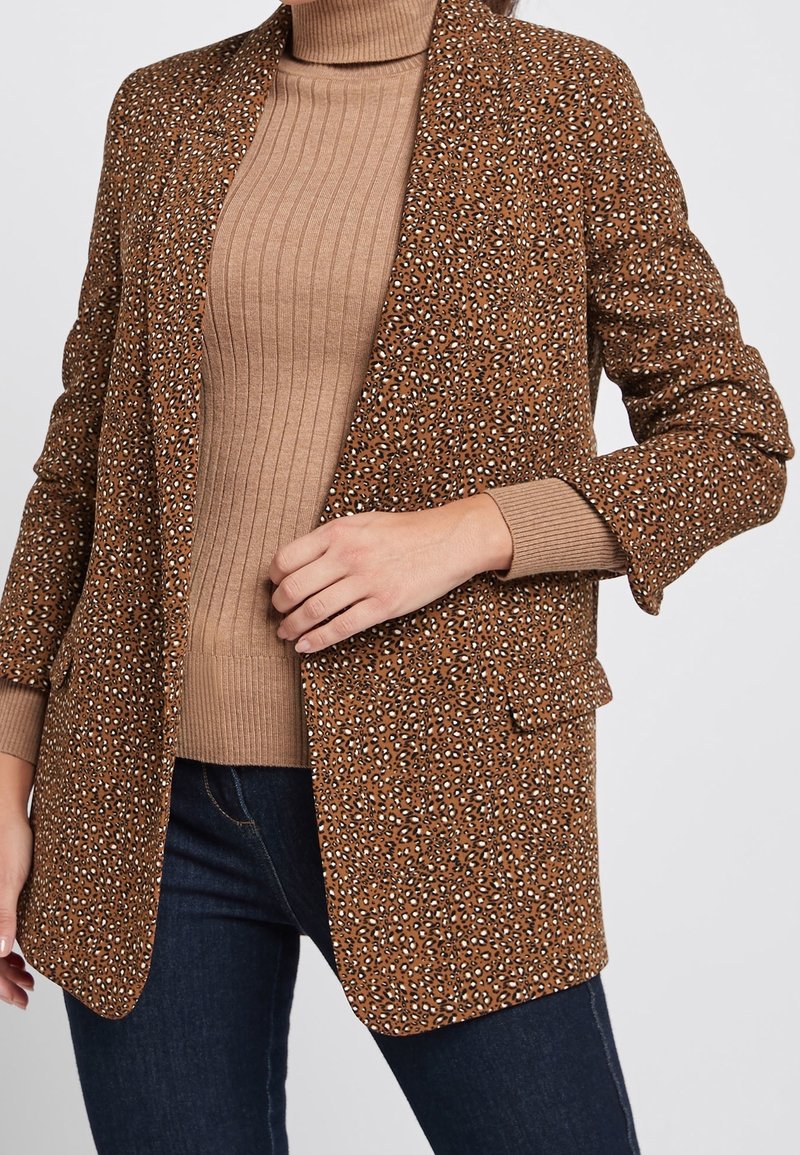 Bruine geblokte blazer met een gestructureerde afwerking, met lichte en donkere vlekken, en een klassiek notch lapel. Draagt over een geribbelde beige top.