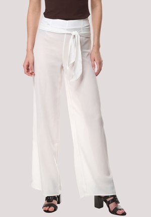 Mujer vistiendo pantalones de pierna ancha blancos con un cinturón atado en el frente, combinados con sandalias de tacón negras y una blusa sin mangas oscura.