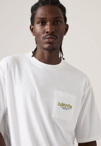 Άσπρο βαμβακερό T-shirt με κοντά μανίκια, διαθέτει τσέπη στο στήθος και κίτρινο-μπλε λογότυπο «Levi's» στην τσέπη. Ομαλή υφή.