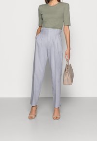 Højtaljede, lyseblå linnedbukser med sidelommer og plisseret front. Sammen med en tætsiddende olivengrøn bomuldsbluse og en beige taske.