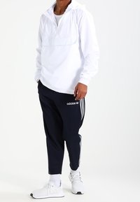 Hvid pulloverjakke med halv lynlås og frontlomme, parret med navy trackbukser med hvide Adidas-striber og logo. Hvide sneakers.
