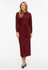 Bordowa, prążkowana sukienka z długim rękawem, dopasowana w talii z paskiem do związania, sięgająca do połowy łydki. Stylizację dopełniają czarne, płaskie buty.