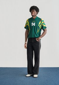 Πράσινο αθλητικό μπλουζάκι με κίτρινες και πράσινες λεπτομέρειες, λευκός αριθμός 94· συνδυασμένο με μαύρο παντελόνι καμπάνα και λευκά αθλητικά παπούτσια.