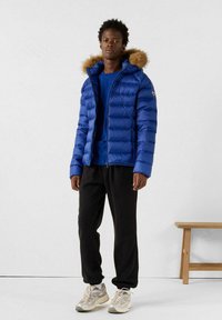 Hombre joven de pie en interiores, vestido con una chaqueta acolchada azul con capucha de piel, camiseta azul, pantalones negros y zapatillas beige, al lado de un banco de madera.