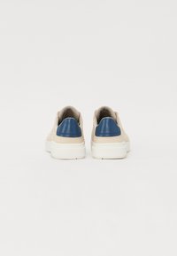 Sapatilhas bege com um detalhe azul no calcanhar, material suave, colarinho almofadado e sola de borracha branca. Design simples e moderno, vista posterior.