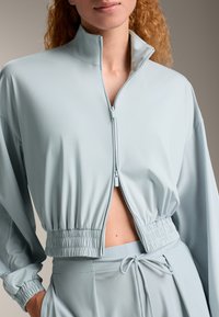 Svijetloplavi skrateni kaput napravljen od glatkog materijala, s uzdignutim ovratnikom i elastičnom donjom rubom. Prednji dio sa zatvaračem i precizno detaljno šivanje.