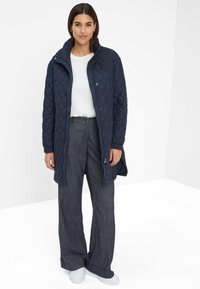 Mørkeblå quilted lang jakke med høj krave og lynlås, parret med grå stribede vide bukser og hvide sneakers.