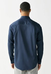 Mężczyzna w ciemnoniebieskiej koszuli z długim rękawem i guzikami, stojący tyłem, w połączeniu z jasnoszarymi spodniami.