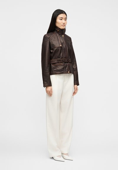 Γυναίκα που στέκεται φορώντας σκούρο καφέ δερμάτινο μπουφάν με ζώνη, λευκό παντελόνι με φαρδιά πόδια και λευκά μυτερά παπούτσια, σε απλό φόντο.