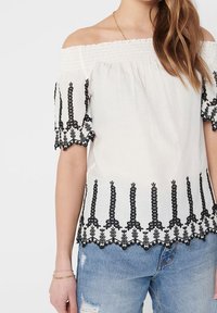 Blouse blanche à épaules dénudées avec broderie noire, présentant un col froncé et des motifs floraux le long des manches et de l'ourlet. Associée à un jean déchiré.