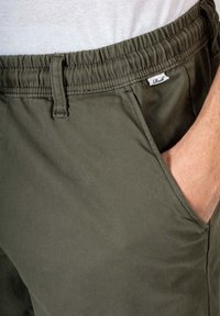 Pantalones cortos de color verde oliva con cintura elástica y cordón, bolsillos laterales y una pequeña etiqueta blanca cosida cerca de la cintura. Tejido suave de mezcla de algodón.